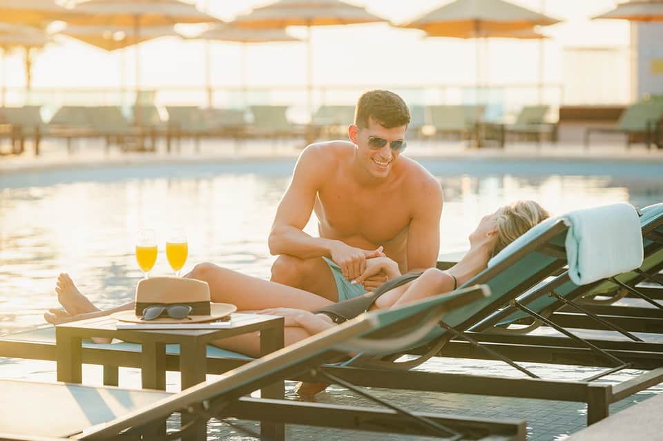 Couple relaxing on lounge chairs by indoor pool with champagne and snacks