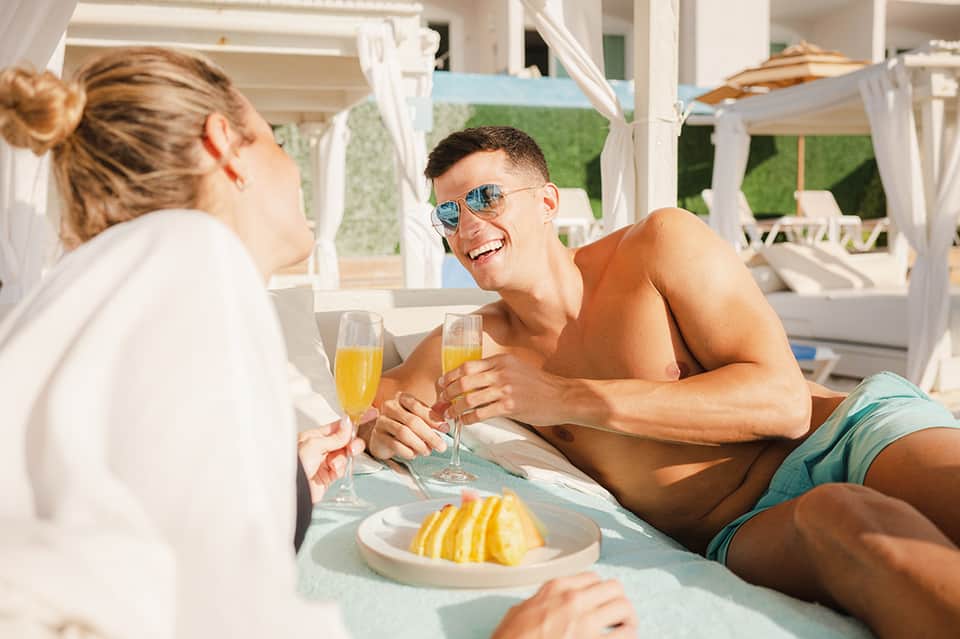 Young couple enjoying tropical drinks and food at beachside cabana with white curtains