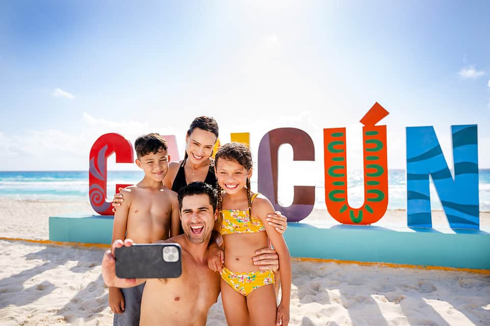 Family of four on beach with large colorful Cancun sign, taking selfie in swimwear