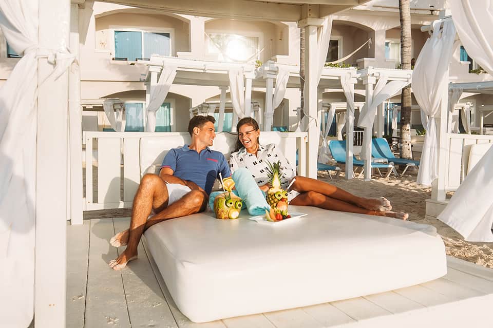 Couple relaxing on white poolside daybed with tropical drinks and beach cabanas in background