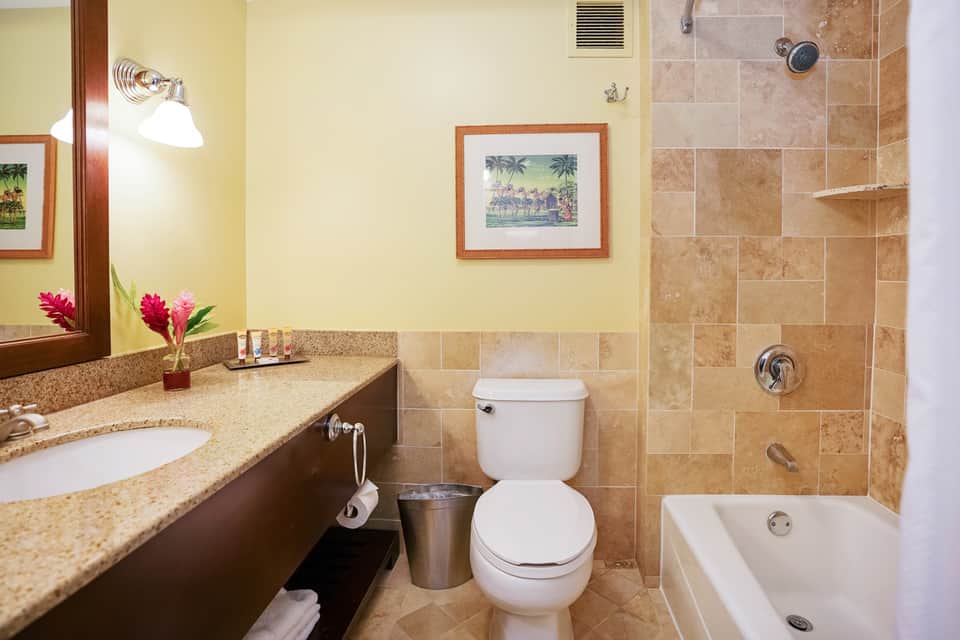 Hotel bathroom with granite vanity, tropical-themed artwork, beige tile shower, and lighting fixtures
