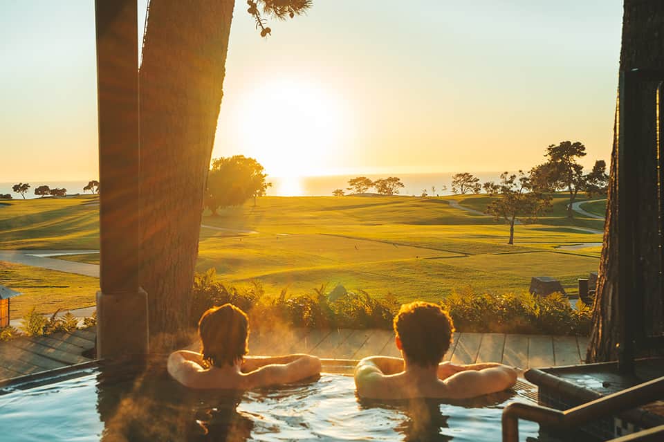 Two people relaxing in outdoor hot tub at sunset overlooking golf course and water views