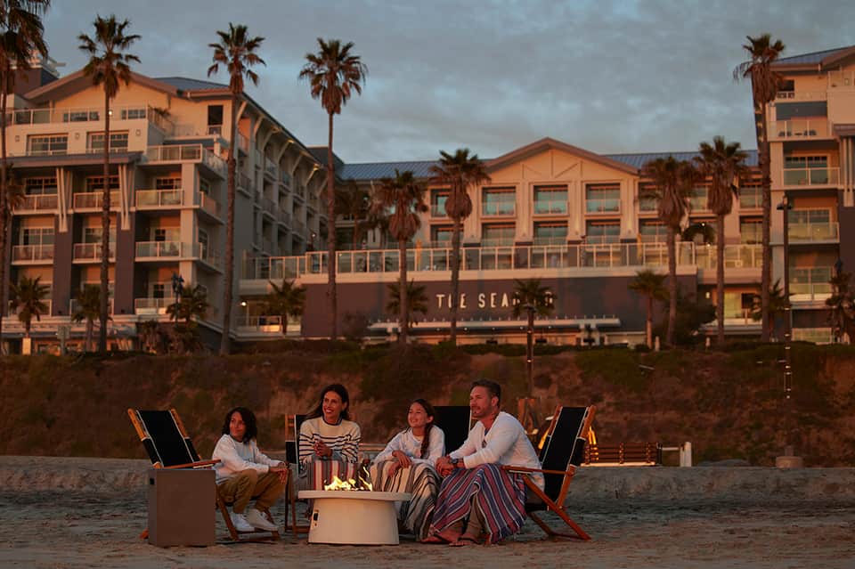 Four people gathered around fire pit on beach at dusk with resort buildings and palm trees in background