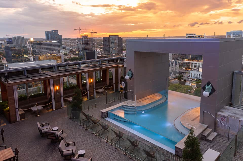 Rooftop pool at sunset with city skyline, lounge chairs, and illuminated blue water features
