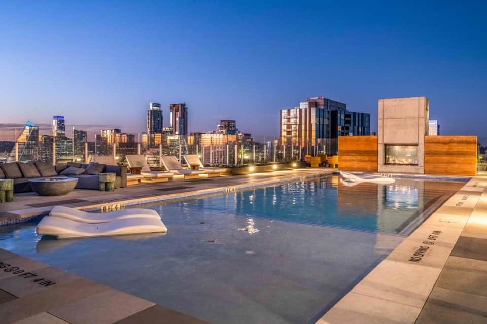 Rooftop pool at dusk with city skyline views, modern lounge chairs, and blue-lit swimming pool