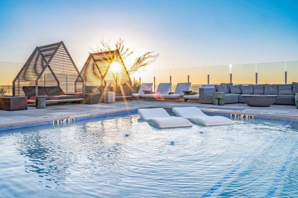 Modern rooftop pool with loungers, geometric shade structures, seating areas, and sunset skyline views