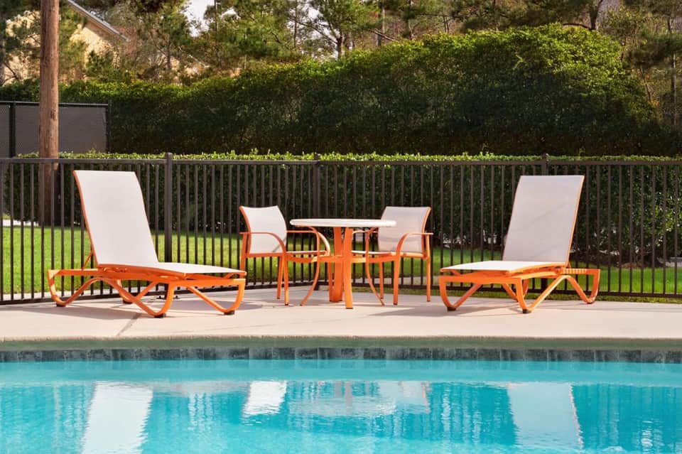 Poolside loungers with orange frames and white cushions beside turquoise water and green hedge privacy fence