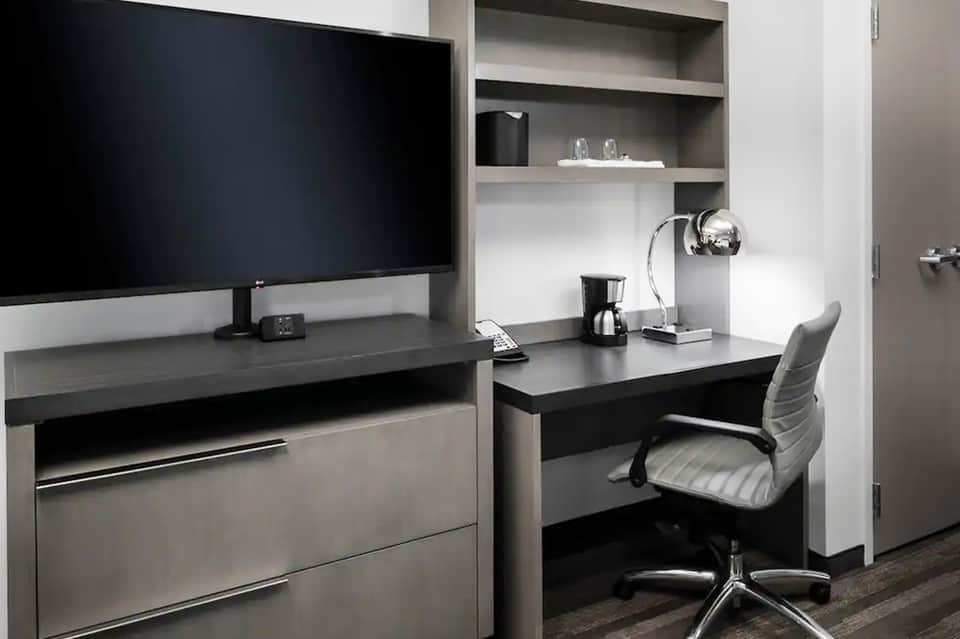 Modern hotel room workspace with black desk, monitor, and built-in shelving unit
