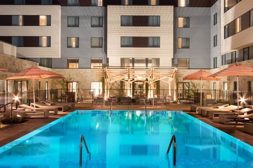Rectangular resort pool at night with blue lighting, umbrellas, lounge chairs, and multi-story hotel building