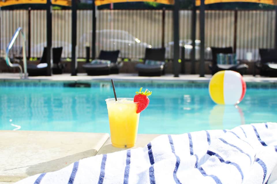 Yellow cocktail with strawberry garnish on pool deck beside striped towel and beach ball