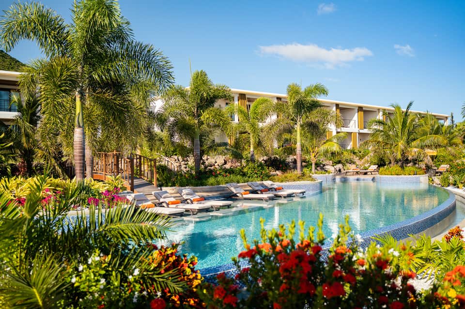 Resort pool with tropical landscaping, colorful flowers, palm trees, and modern hotel buildings