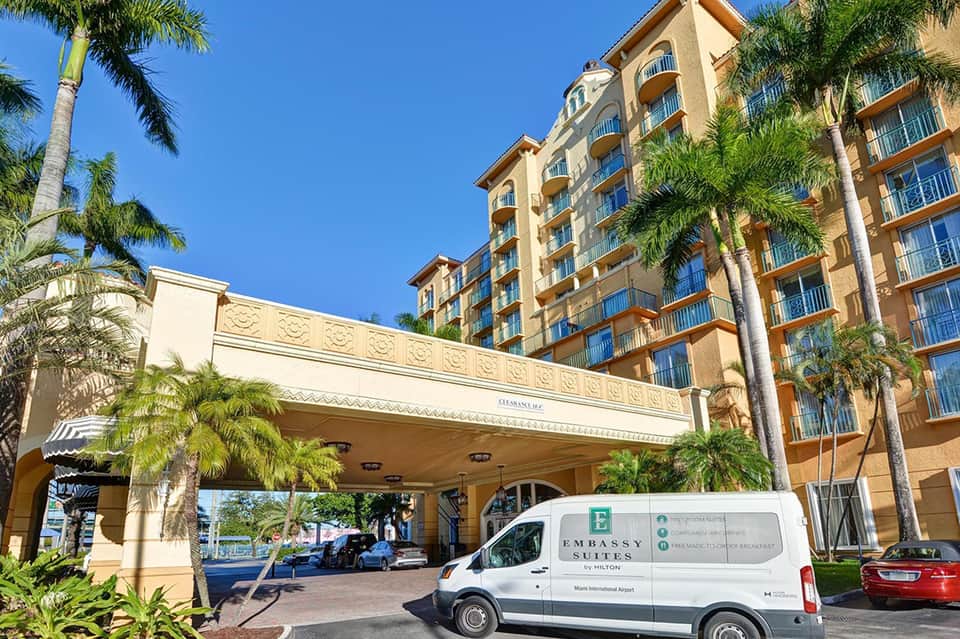 Tropical resort entrance with palm trees, beige architecture, and Embassy Suites shuttle van
