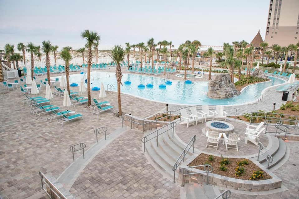 Large resort pool complex with turquoise water, palm trees, lounge chairs, and high-rise hotel building