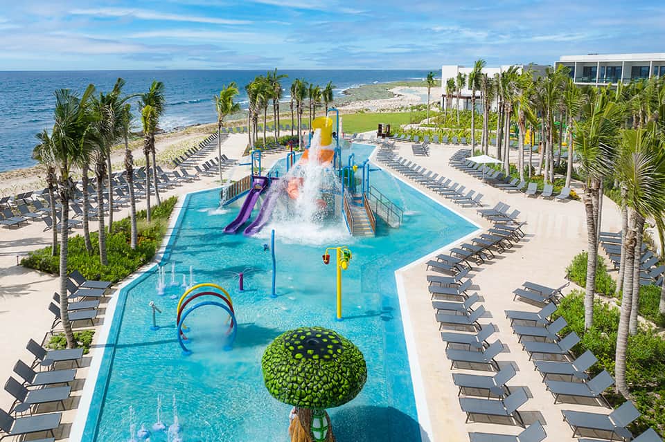 Aerial view of colorful waterpark with slides, shallow pool, palm trees, loungers, and beach beyond