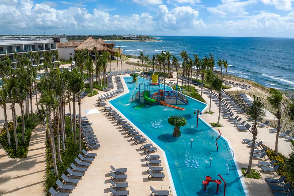 Aerial view of beachfront resort with pools, playground, and ocean horizon