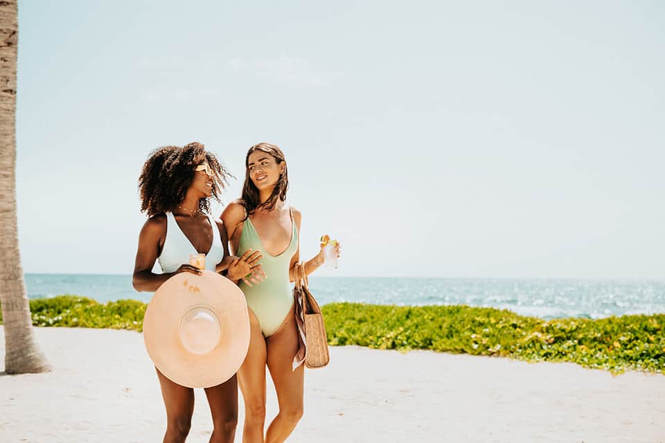 Two women in swimwear on beach with ocean view, one holding a tan hat and woven bag