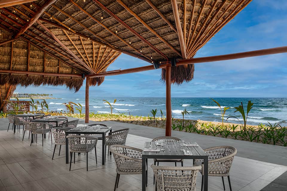 Beachfront dining area with thatched wooden pergola, modern chairs, ocean waves, and tropical vegetation