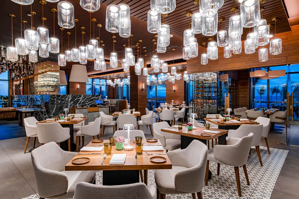 Elegant waterfront restaurant with wooden ceiling, pendant lighting, and expansive windows overlooking water at dusk
