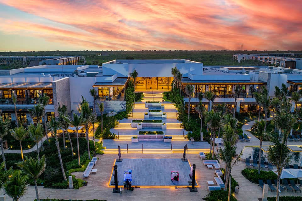 Aerial dusk view of modern resort with illuminated courtyard, palm trees, and colorful sunset sky