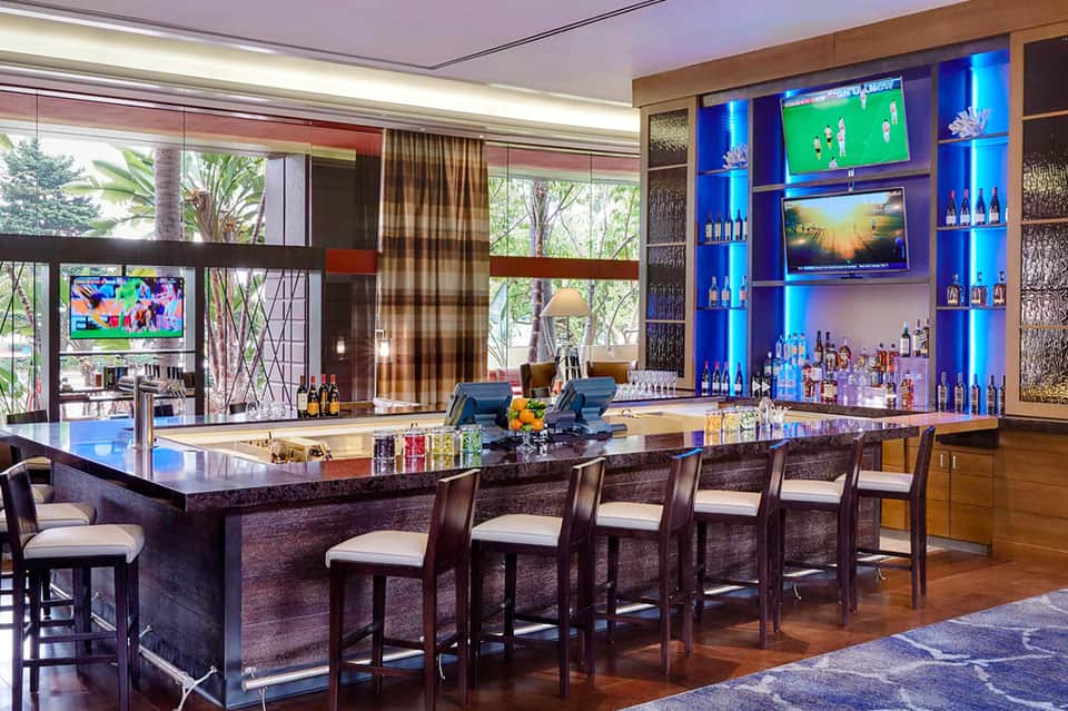 Modern bar lounge with curved black counter, blue-lit shelving, multiple screens, and bamboo garden views