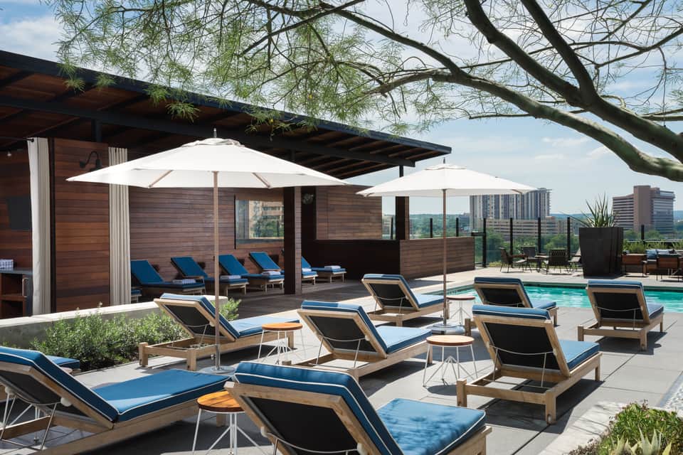 Rooftop pool deck with blue loungers, white umbrellas, wood cabana, and city skyline in background