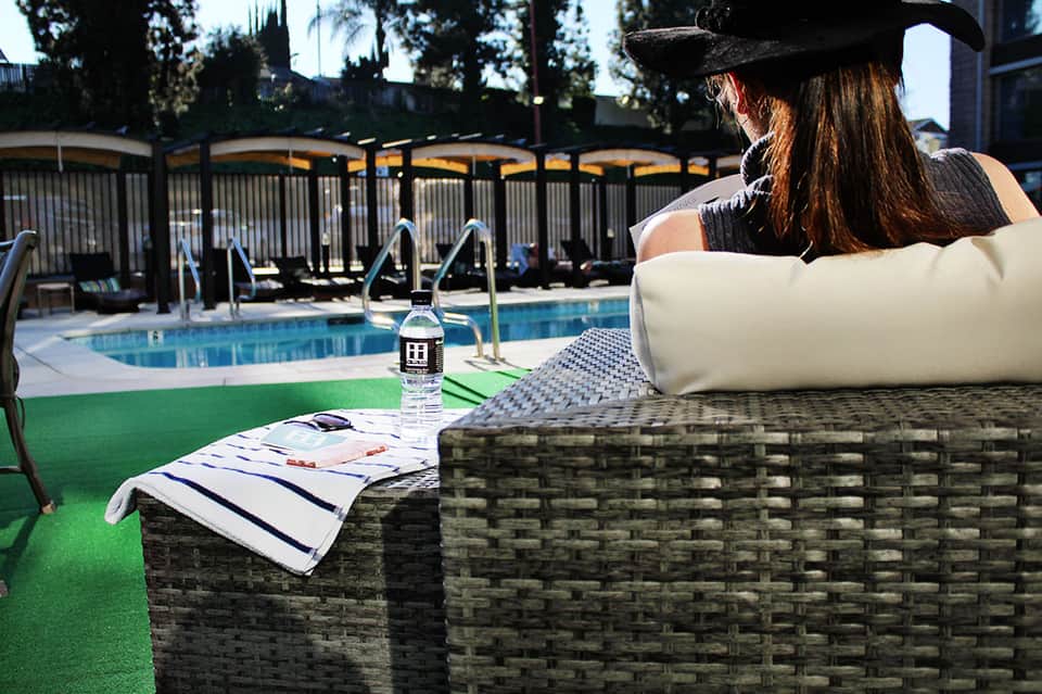 Woman in hat relaxing in lounge chair by resort pool with fence and palm trees