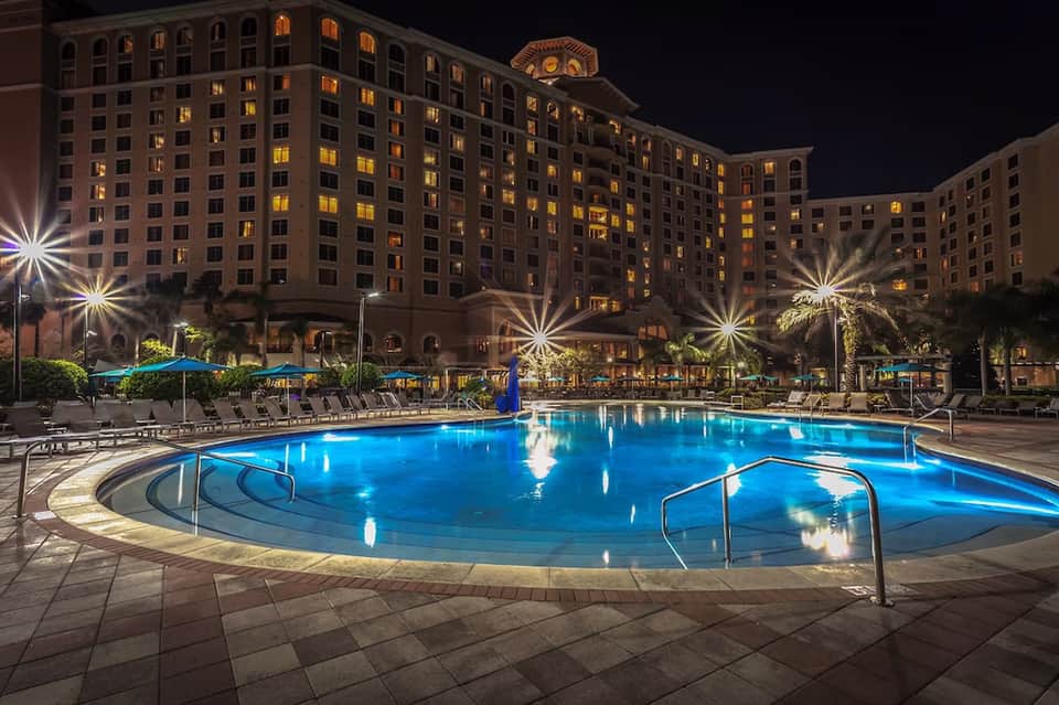 Illuminated resort pool at night with multi-story hotel building, palm trees, and blue umbrellas