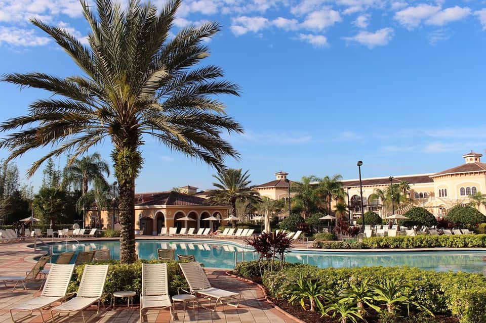 Resort pool area with palm trees, lounge chairs, and Mediterranean-style yellow buildings