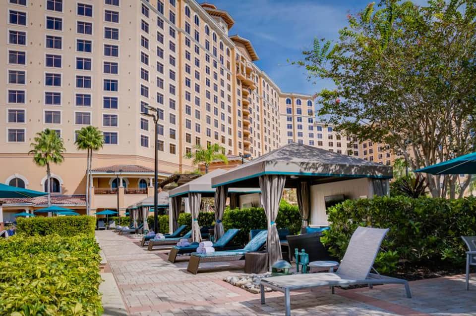 Resort pool deck with lounge chairs, cabanas, palm trees, and multi-story hotel building