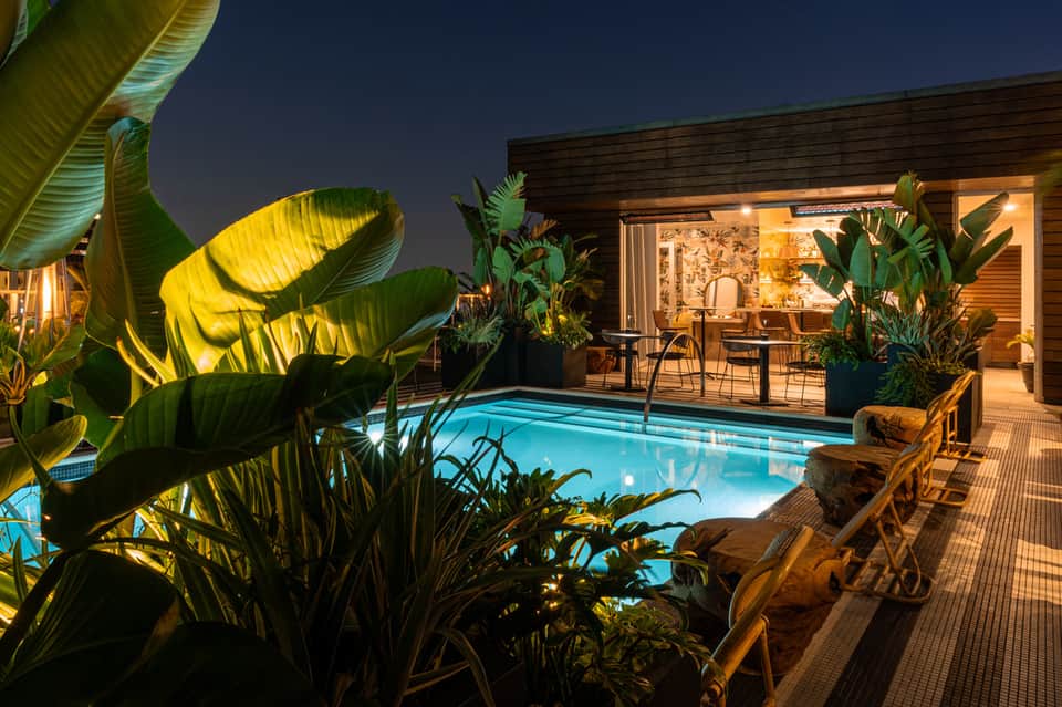 Nighttime resort pool with tropical plants, lounge chairs, and illuminated wooden pavilion with indoor seating visible