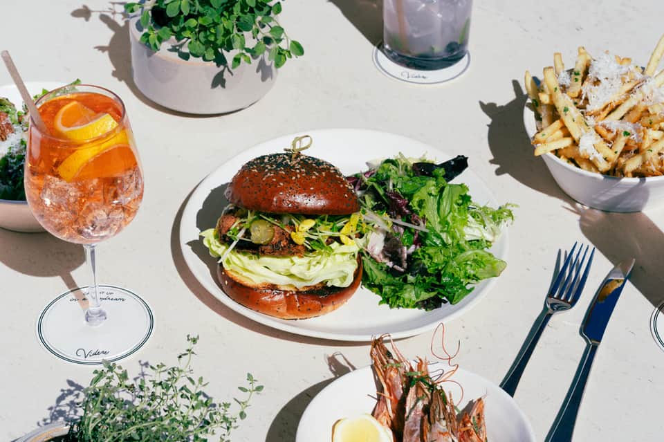 Outdoor dining table with gourmet burger, fries, and cocktail on sunny terrace with potted plants