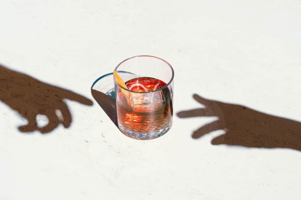Glass of amber cocktail with shadow of hands on white surface in sunlight