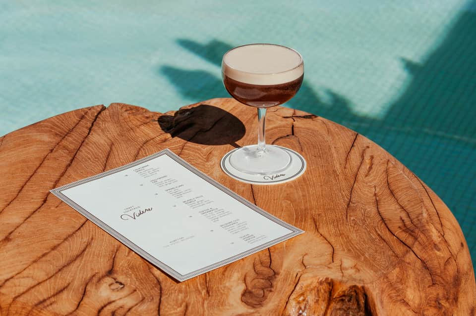 Elegant wooden table with cocktail menu and espresso martini overlooking turquoise pool waters