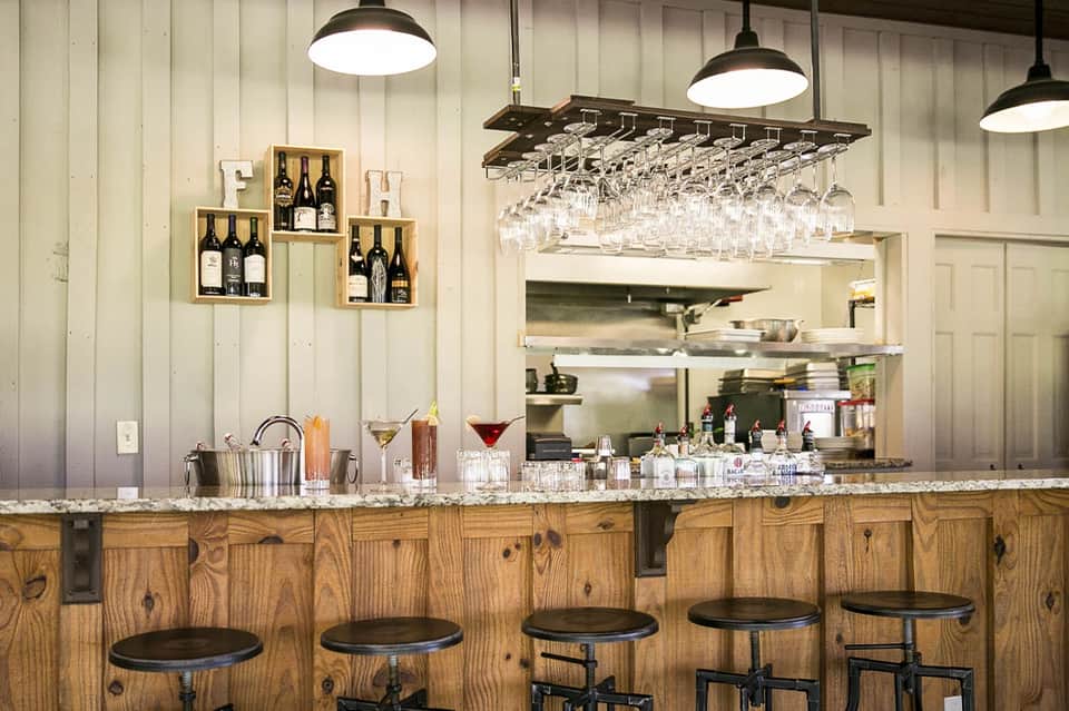 Modern wine bar with wooden counter, black bar stools, pendant lights, and wine bottle displays on white shiplap walls