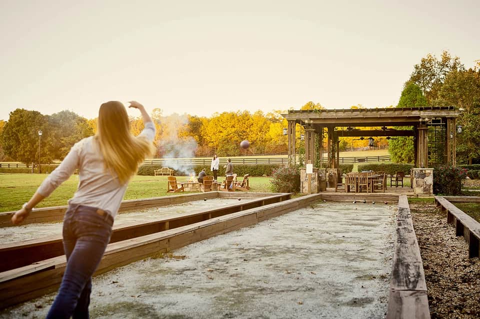 Woman playing cornhole at a rustic outdoor pavilion with fire pit and pastoral views
