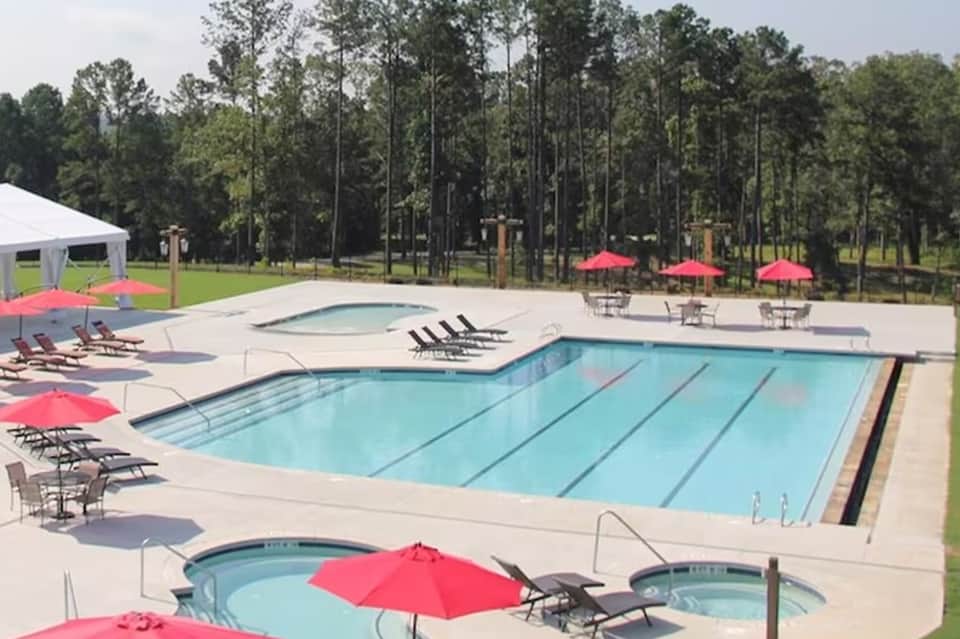 Resort swimming pool with lap lanes, hot tubs, and pink umbrellas surrounded by lounge chairs and tall pine trees
