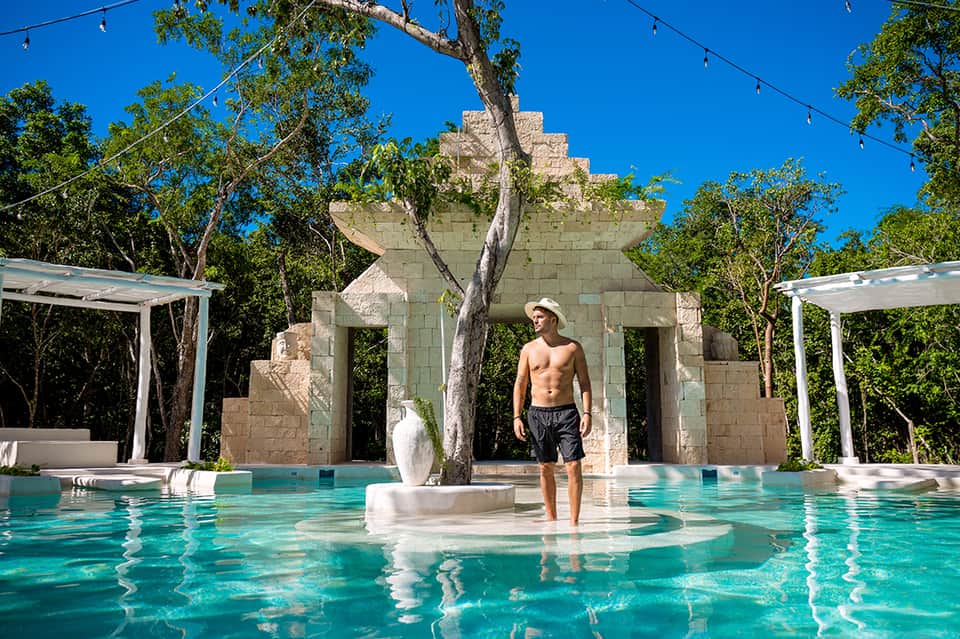 Turquoise pool with stone archway and man standing in shallow water surrounded by tropical trees