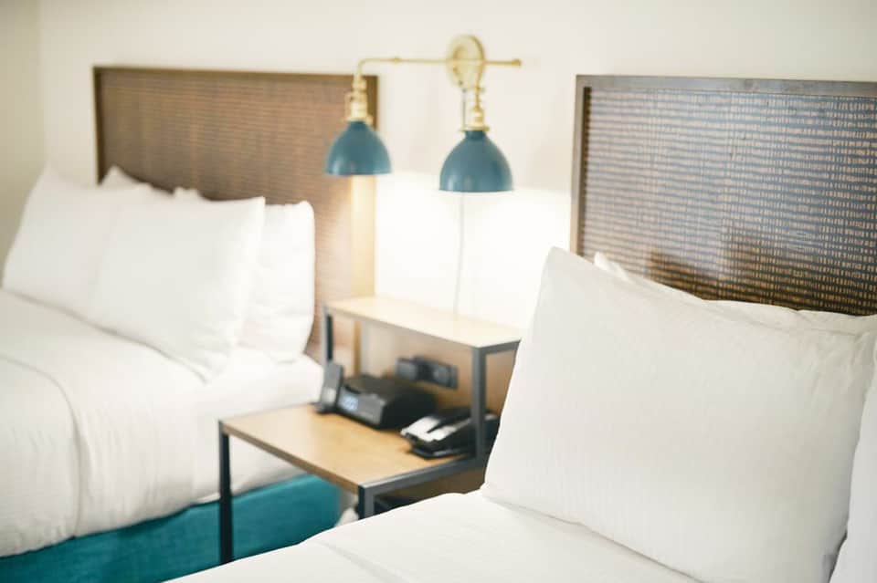 Modern hotel bedroom with two queen beds, white bedding, teal accent wall, and blue pendant lighting fixtures