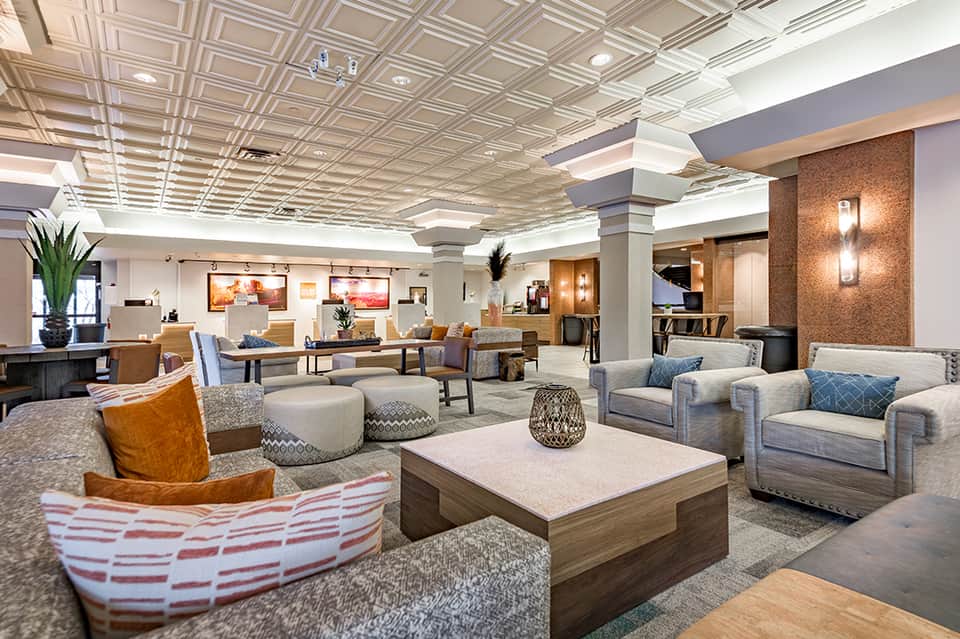 Modern hotel lobby with geometric ceiling, comfortable seating areas, neutral tones, and decorative plants