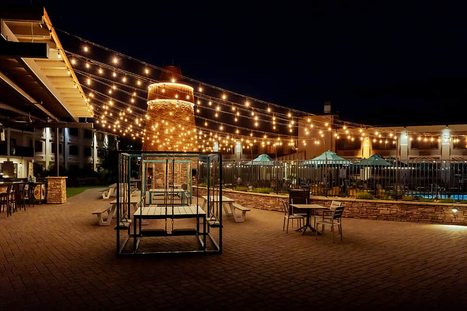 Illuminated outdoor patio at night with string lights, gazebo structures, and brick fireplace tower