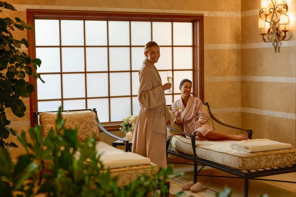 Two women in spa robes relaxing in a luxurious spa room with large windows and beige tones