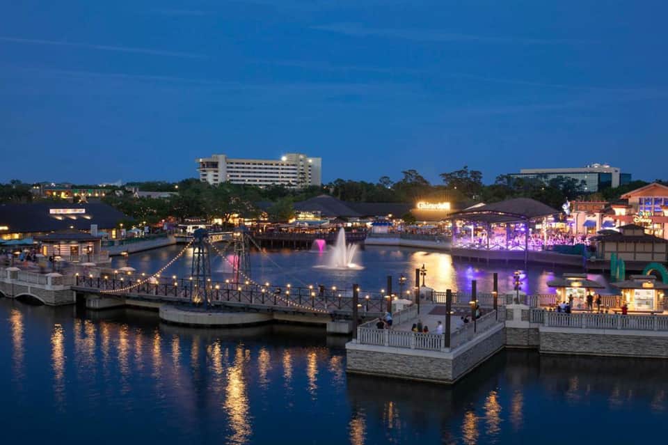 Illuminated waterfront entertainment complex at dusk with fountain, bridges, and colorful purple-lit buildings