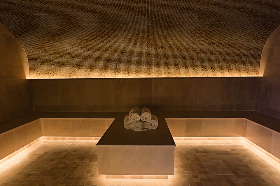 Luxurious spa treatment room with mosaic tile ceiling, heated stone basin, and rolled towels on dark stone platform