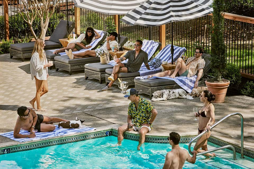 Resort pool deck with guests relaxing on lounge chairs under striped umbrellas and tropical landscaping