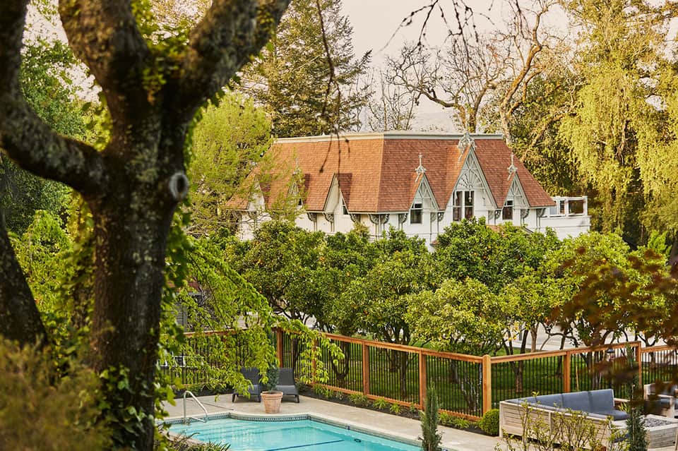 Elegant white manor with terracotta roof nestled among tall trees, view from pool area with wooden deck