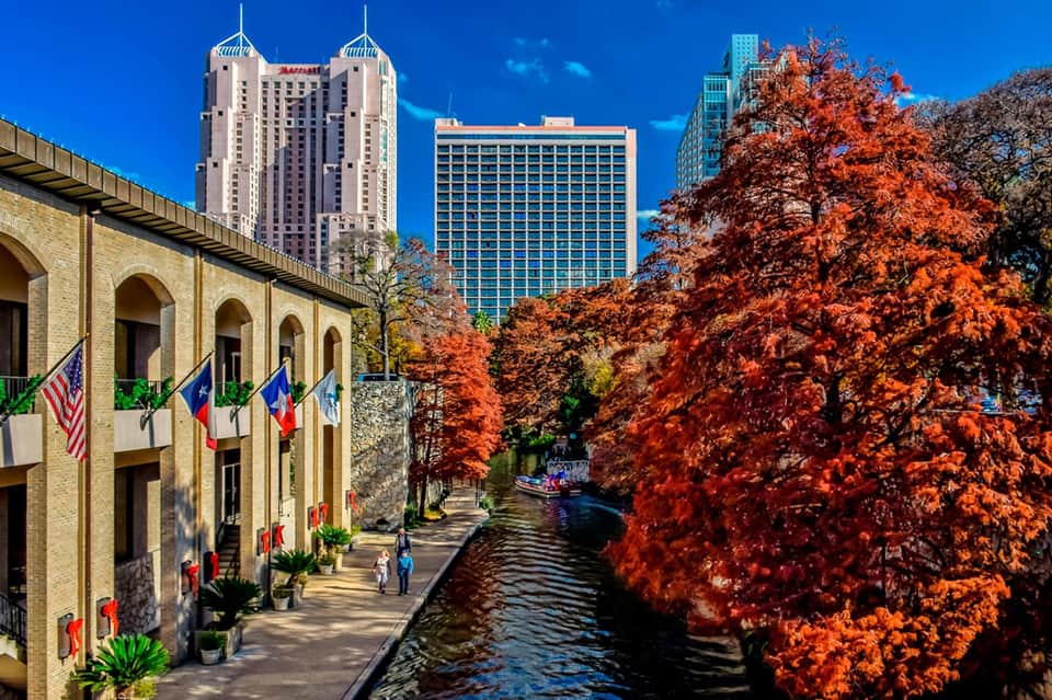 Scenic waterfront promenade with historic stone building, modern skyscrapers, and vibrant red autumn foliage