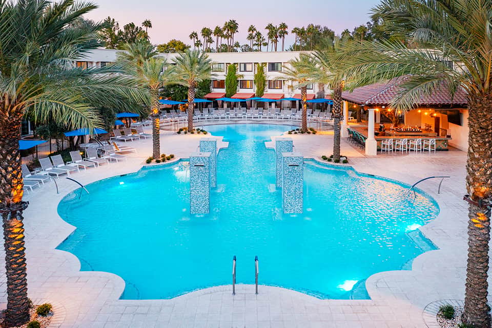 Luxury resort pool with turquoise water, palm trees, and multi-story hotel building with blue umbrellas