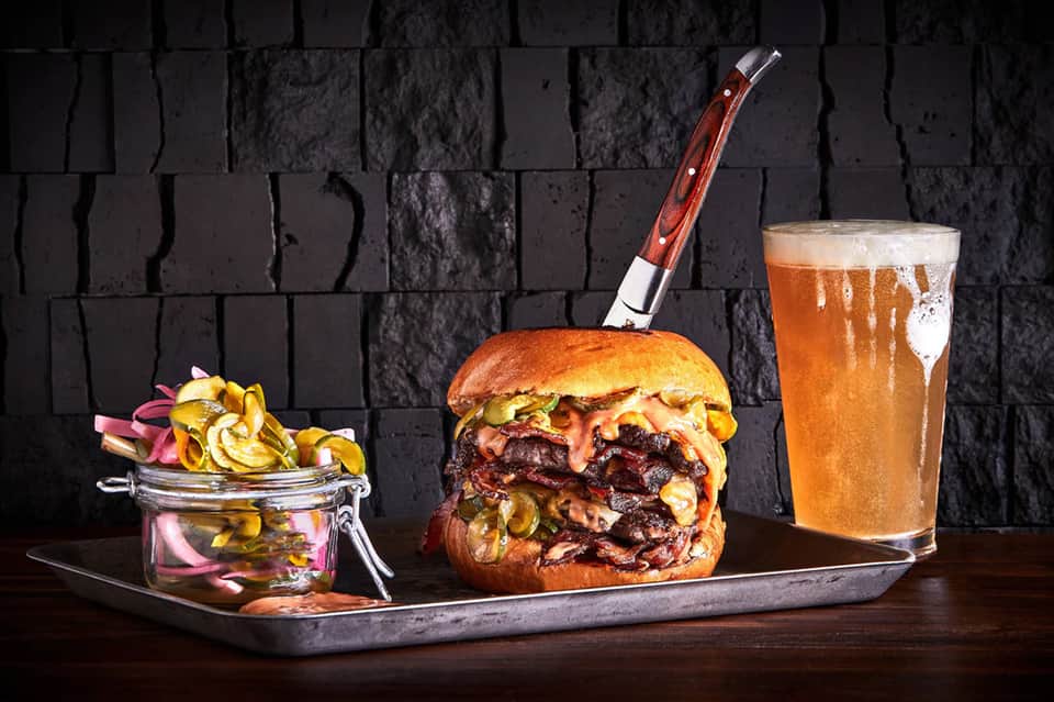 Gourmet burger with caramelized onions, pickles, fries, and craft beer on dark slate background