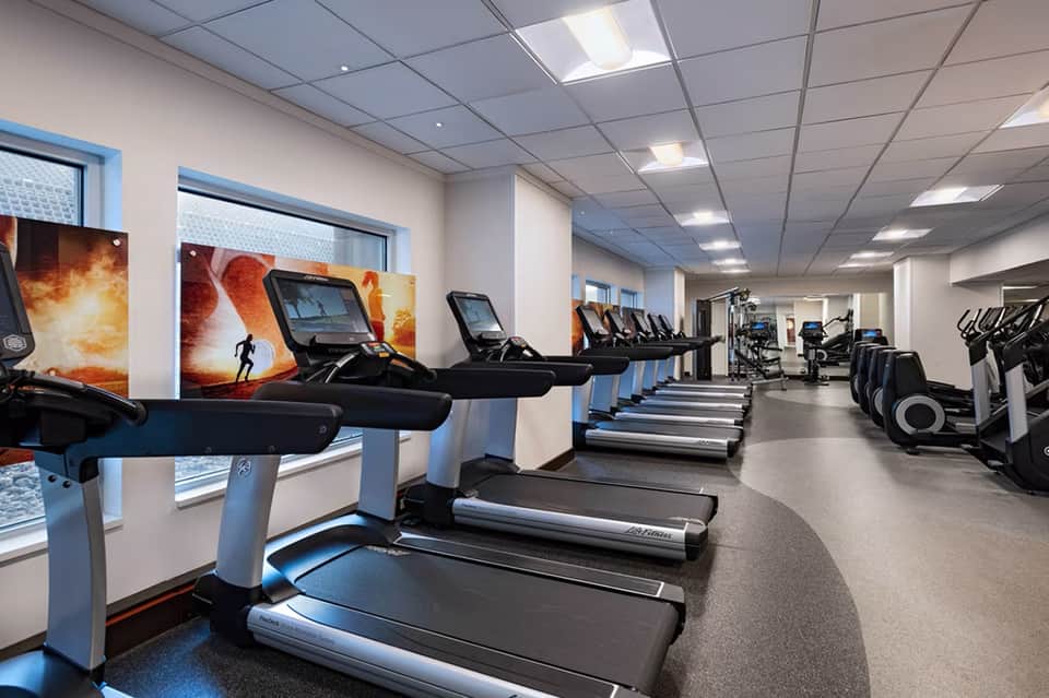 Modern fitness center with rows of treadmills, stationary bikes, and exercise equipment along windowed walls