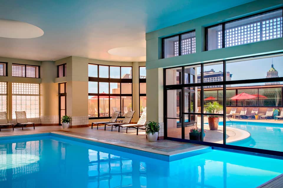 Indoor swimming pool with large windows overlooking an outdoor pool and city skyline